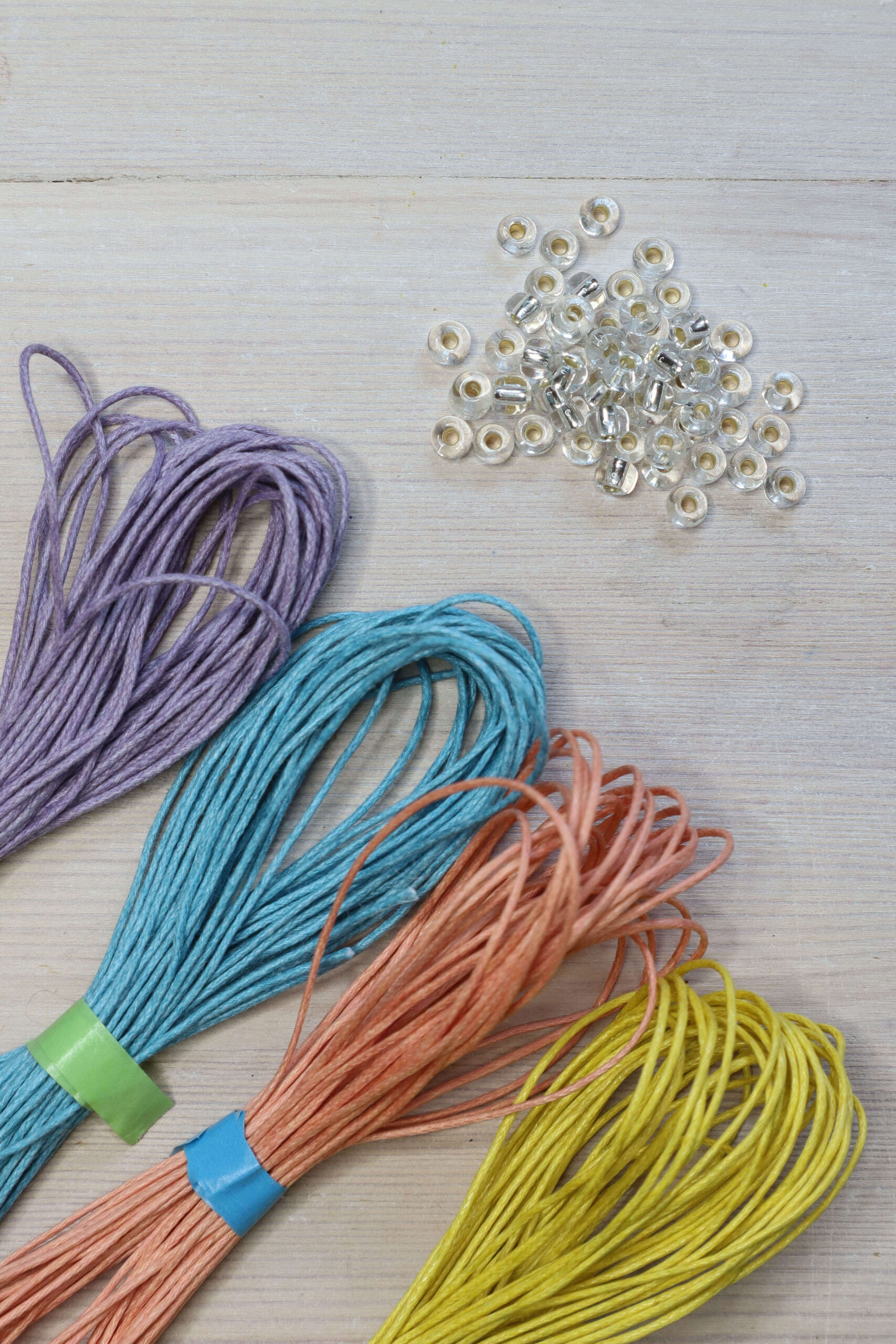 Beaded macrame bracelet kit contents, showing lilac, light blue, peach and yellow cotton cords with shiny clear beads