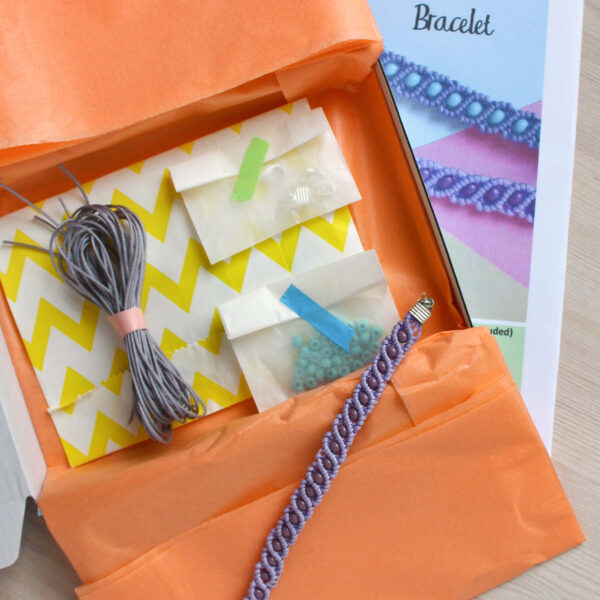 beaded macrame bracelet kit image, showing the finished bracelet in lilac and purple , with the open box containing all the materials and instruction booklet