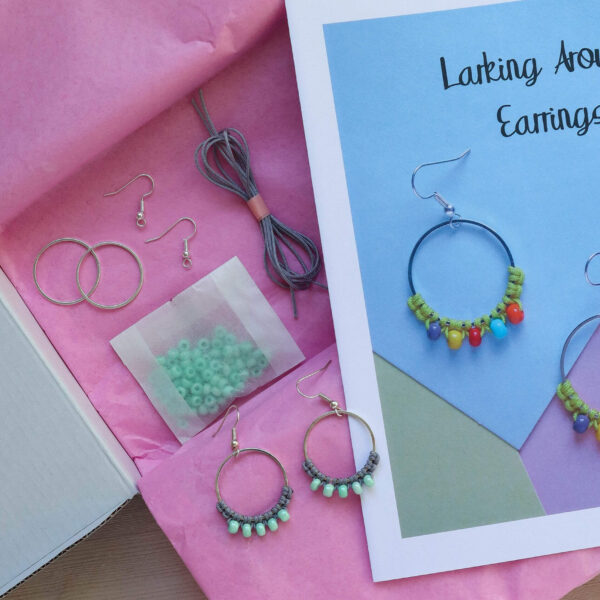 colourful macrame earring kit image showing the open aqua blue box with pink tissue paper and the materials laid out inside along with the all colour instruction booklet and a finsihed pair of grey and mint earrings