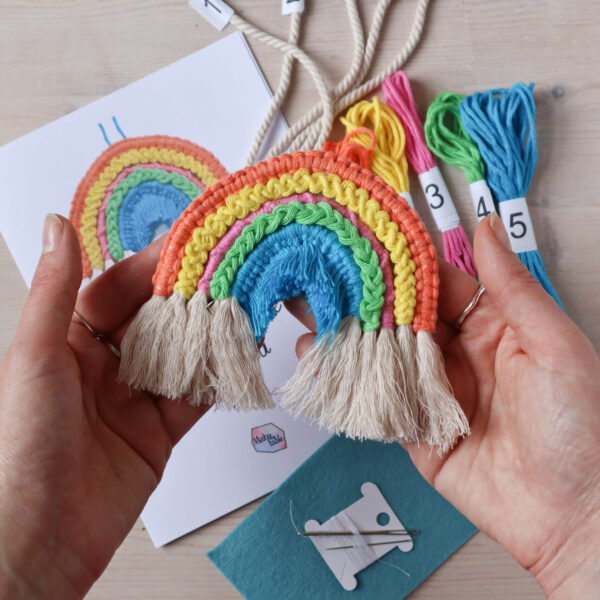 Macrame rope rainbow kit image, showing a finished being held in the hands of a white woman, with the instruction booklet and materials from the kit in the background