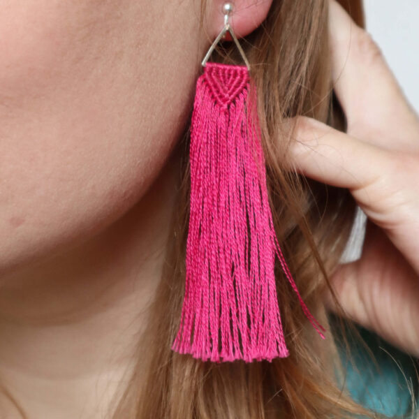 Macrame tassel earrings kit image showinf a white woman wearing a finished pair of knotted earrings in fuchsia pink, with a triangle connector and long fringe that comes just below her chin