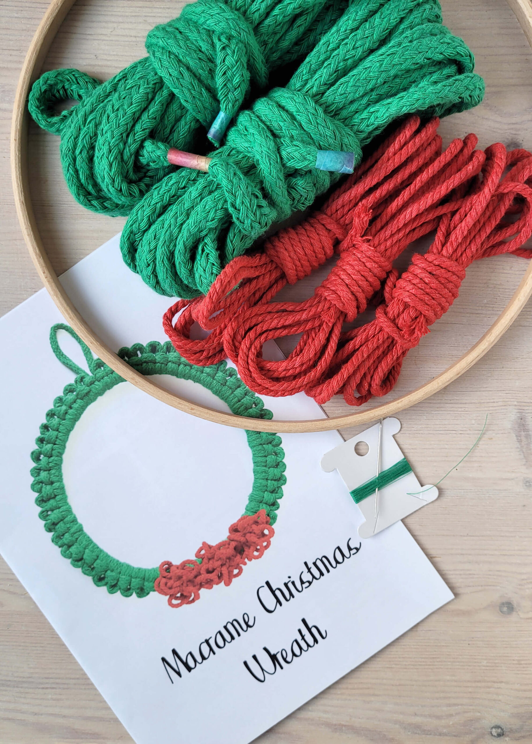 macrame Christmas wreath kit contents, showing instructions, wooden hoop, green and red cord and needle and thread
