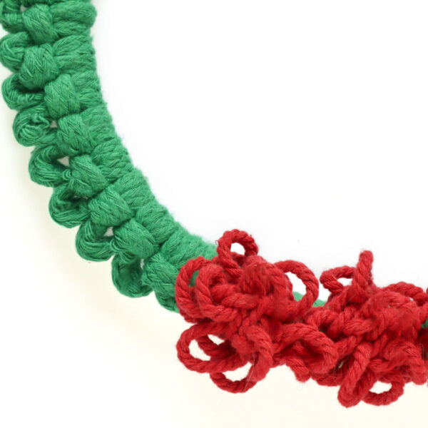 Close up of a macrame Christmas wreath with green cord and bright red pom pom berries on a white wall
