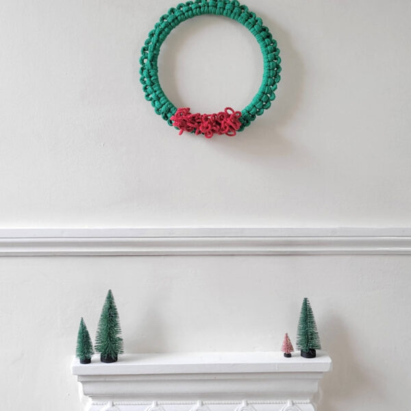 macrame Christmas wreath with green cord and bright red pom pom berries on a white wall above a fireplace