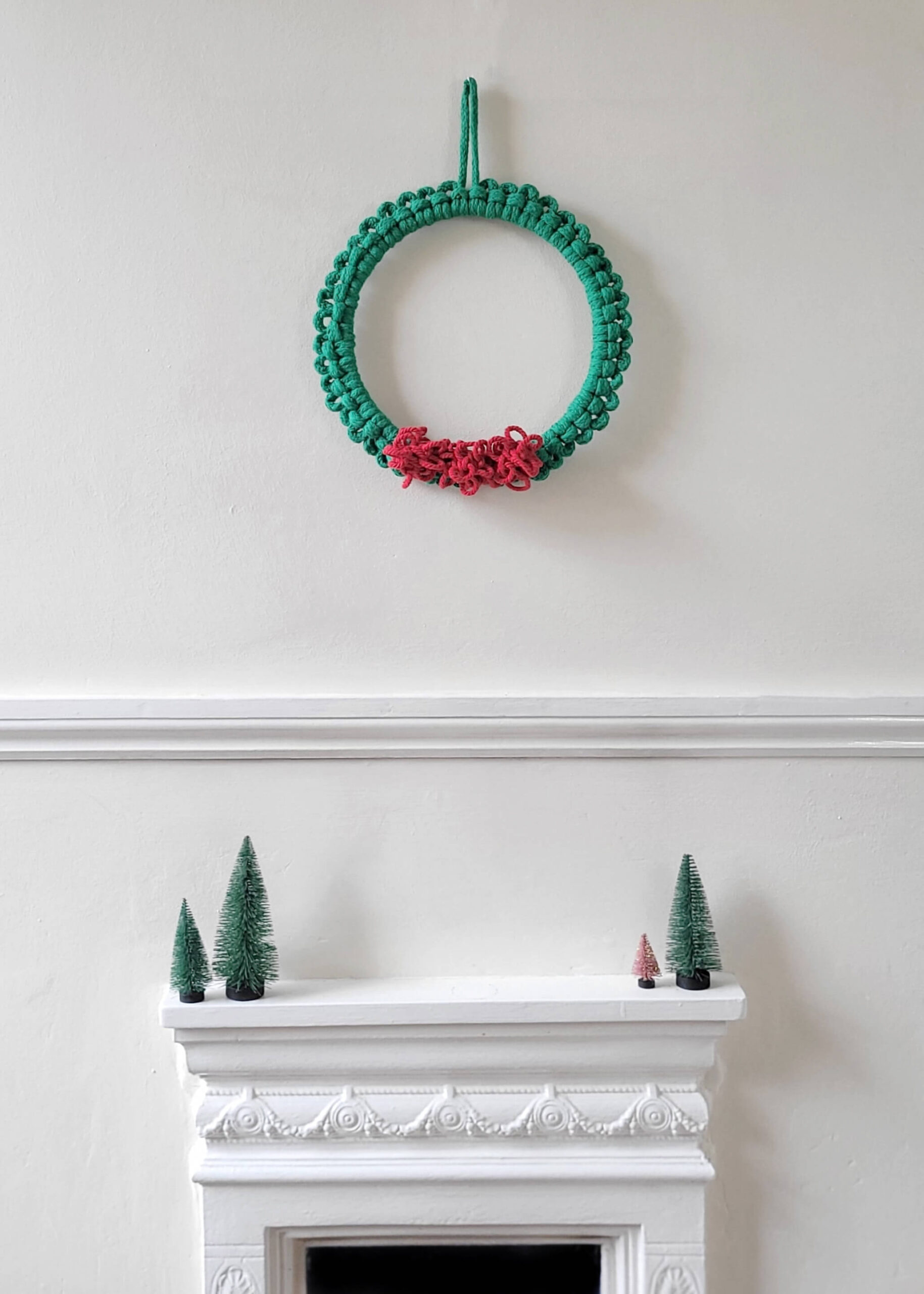 macrame Christmas wreath with green cord and bright red pom pom berries on a white wall above a fireplace