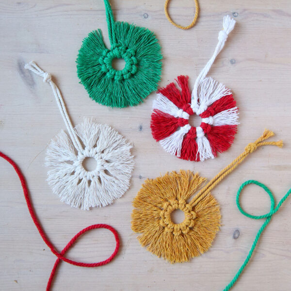 four macrame christmas decorations in green. whire, mustard and a striped white and red, on a wooden background. They are circular with a fringe