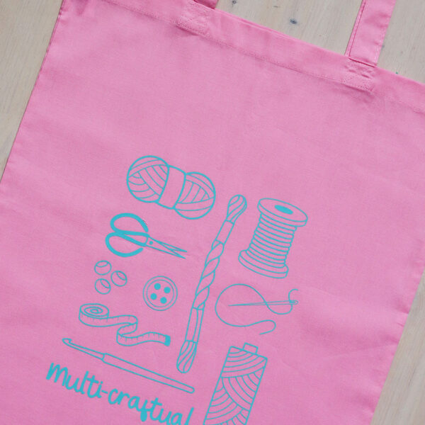 Colourful tote bag, pink with teal print of craft items like macmrae cord, yarn, embroidery thread, sewing thread and tools and the word multicraftual