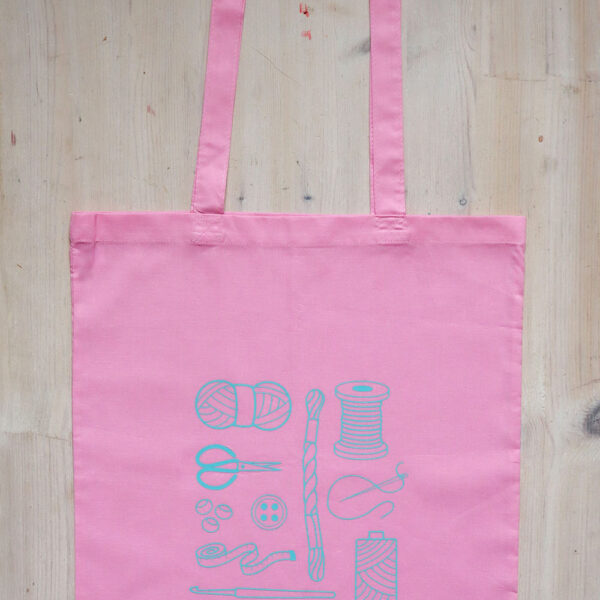 Colourful tote bag, pink with teal print of craft items like macmrae cord, yarn, embroidery thread, sewing thread and tools and the word multicraftual