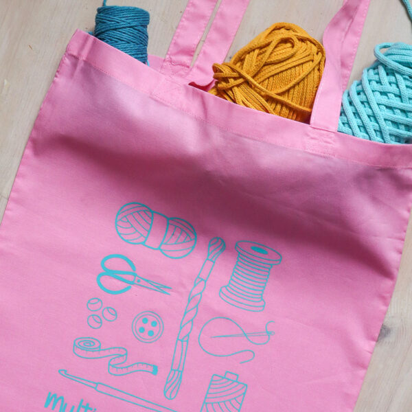 Colourful tote bag, pink with teal print of craft items like macmrae cord, yarn, embroidery thread, sewing thread and tools and the word multicraftual