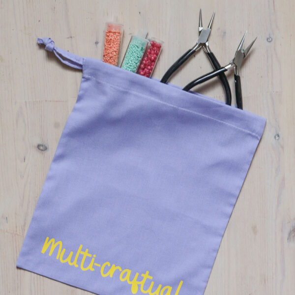 Colourful cotton project bag, pink with teal print of the word multicraftual. There are jewellery making pliers and beads coming out of the bag.