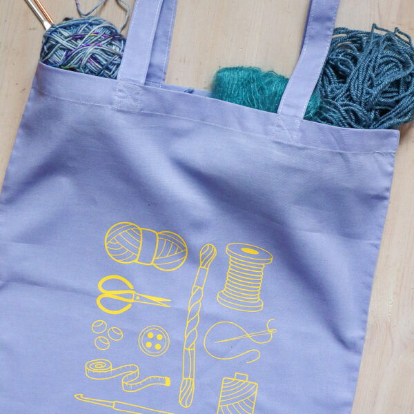 Colourful tote bag, lilac with yellow print of craft items like macmrae cord, yarn, embroidery thread, sewing thread and tools and the word multicraftual