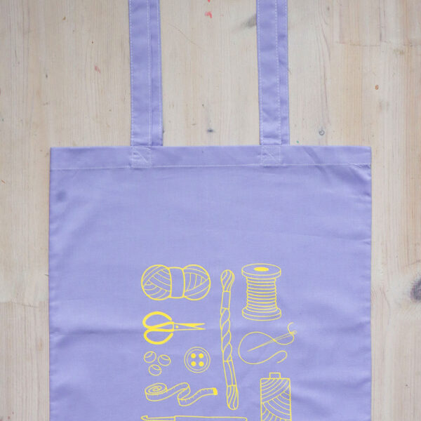 Colourful tote bag, lilac with yellow print of craft items like macmrae cord, yarn, embroidery thread, sewing thread and tools and the word multicraftual