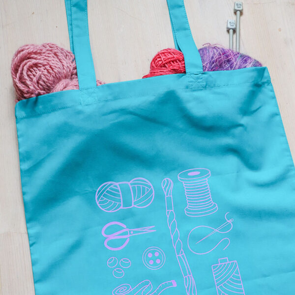 Colourful tote bag, teal with lilac print of craft items like macmrae cord, yarn, embroidery thread, sewing thread and tools and the word multicraftual