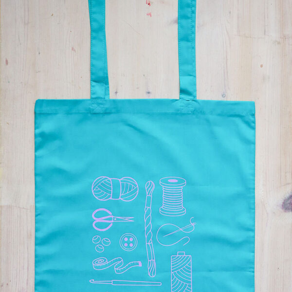 Colourful tote bag, teal with lilac print of craft items like macmrae cord, yarn, embroidery thread, sewing thread and tools and the word multicraftual