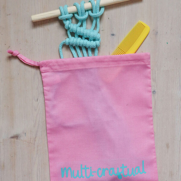 Colourful cotton project bag, pink with teal print of the word multicraftual. There are rolls of cottom cord and a macrame hanging coming out of the bag
