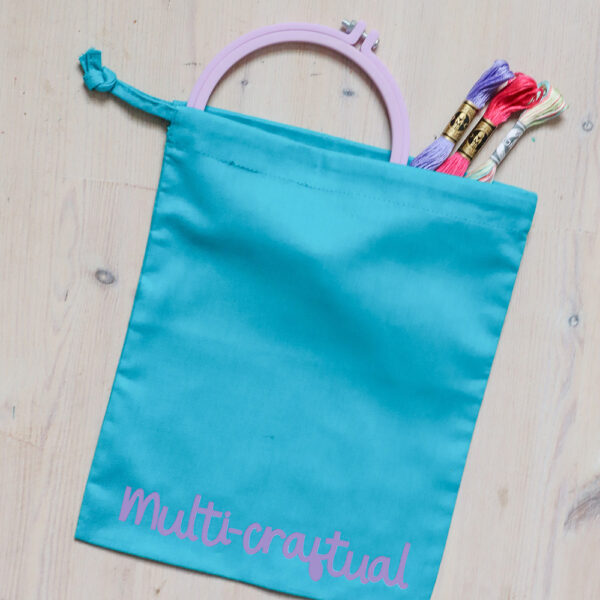 Colourful cotton project bag, teal with a lilac print of the word multicraftual. There is an embroidery hoop and some thread coming out of the bag