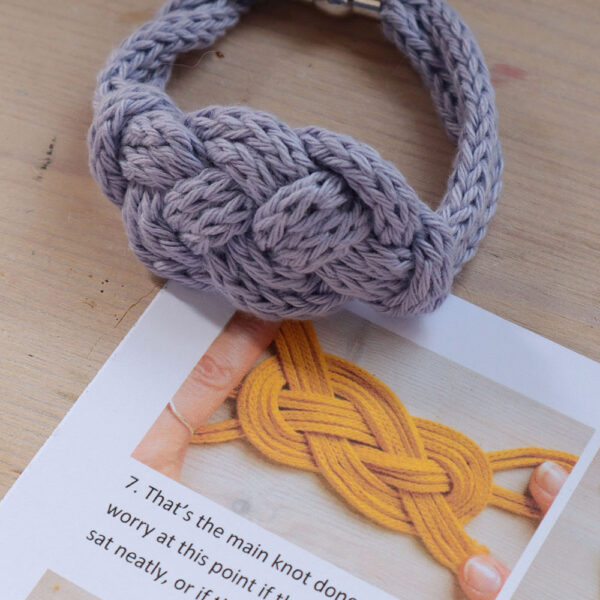 Close up of a printed pattern for the Carrick Cuff, a macrame bracelet pattern sits on a wooden background with a completed purple example