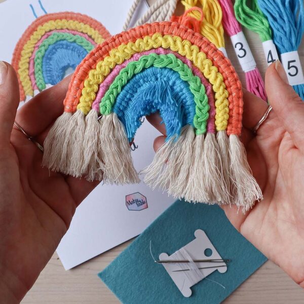 hands holding a completed macrame rainbow, with the materials in the background