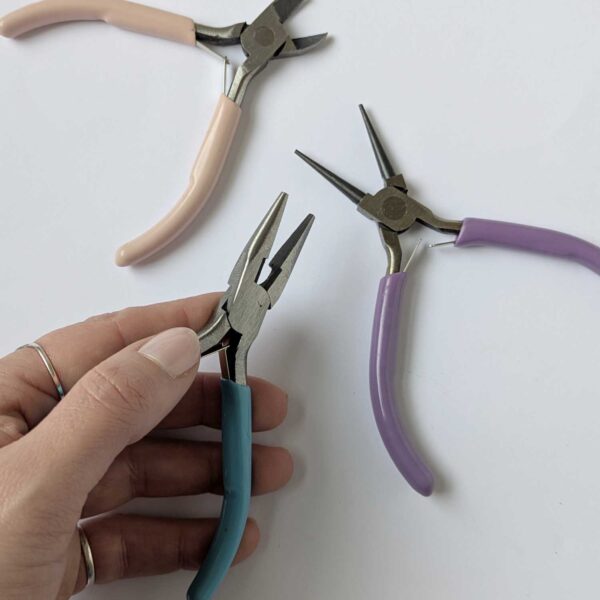 hand holding a pair of chainnose pliers with a blue handle, there are cutter with a pink handle and roundnose pliers with a purple handle in the background