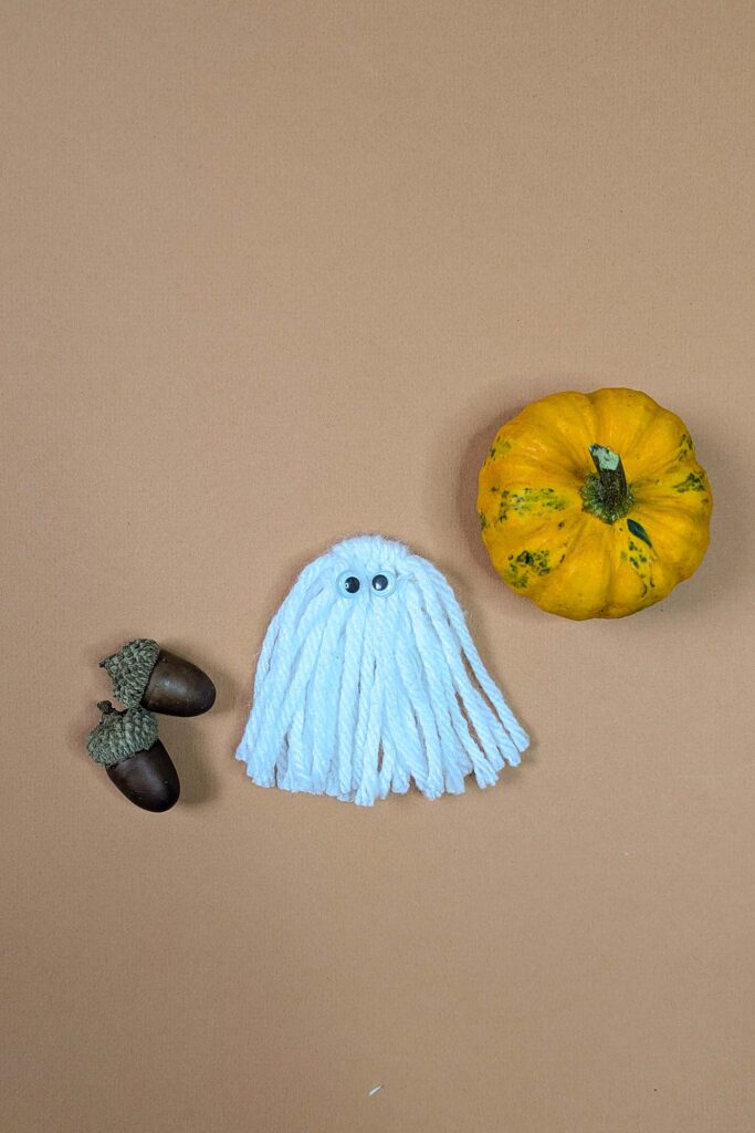 Small macrame ghost made with aran / worsted weight yarn and a 25mm d-ring. It has googly eyes and is on peach background with a pumpkin and acorns
