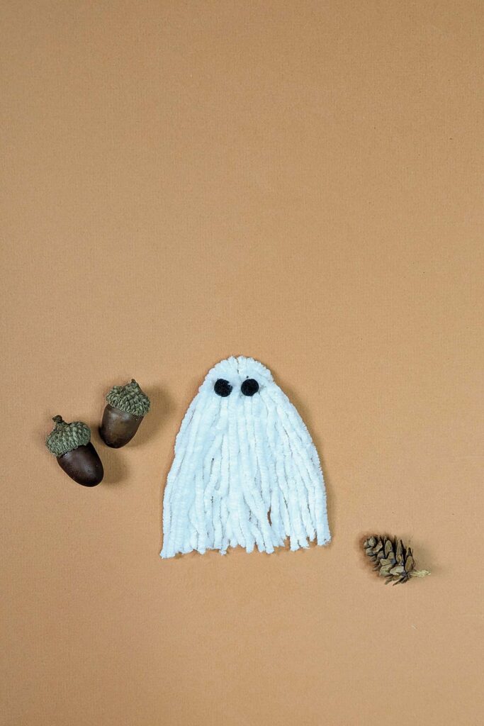 Small macrame ghost made with 25mm d-ring and white DK weight chenille yarn. It has black pom pom eyes and is on a peach background with acorns and a pine cone