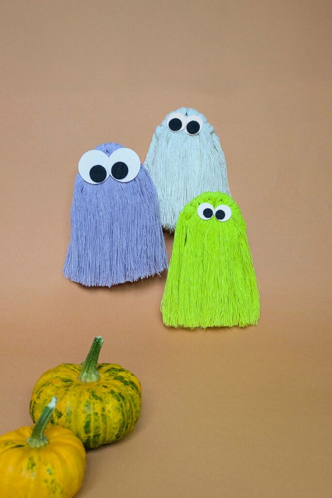 Three macrame ghost, one in white, one in lilac and one in neon green cord! They each have different sizes of felt googly eyes. There are two pumpkins in the foreground on a peach colour background
