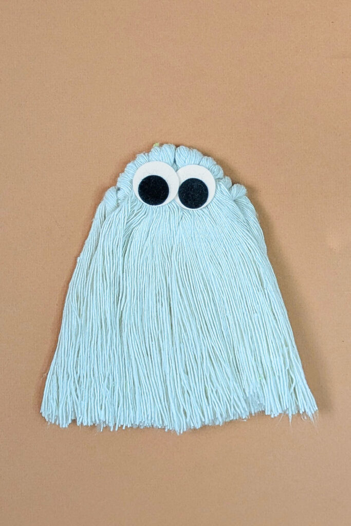 A completed macrame ghost in white cords with felt eyes on a peach background